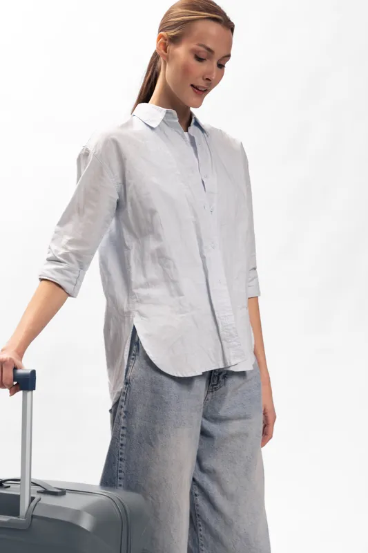 Young woman in casual shirt and jeans stands holding suitcase handle, looking down. Concept of travel, lifestyle, and preparation for trip in bright studio setting.