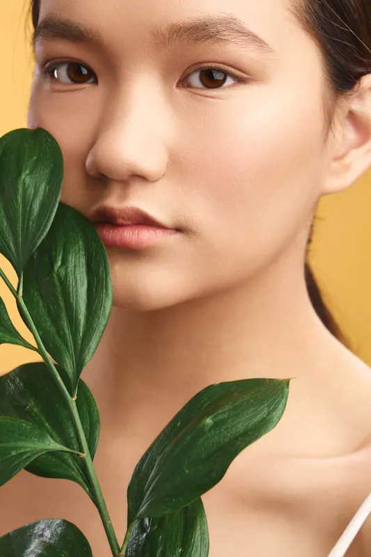 Asian woman gazes softly at the camera in a studio portrait with lush green leaves and a warm backdrop, creating a calm, fresh mood suitable for beauty, lifestyle or wellness topics.