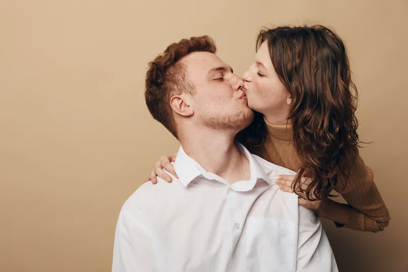 Young couple shares a tender kiss with eyes closed, woman embracing man from behind. Intimate moment of love and affection captured on neutral beige background.