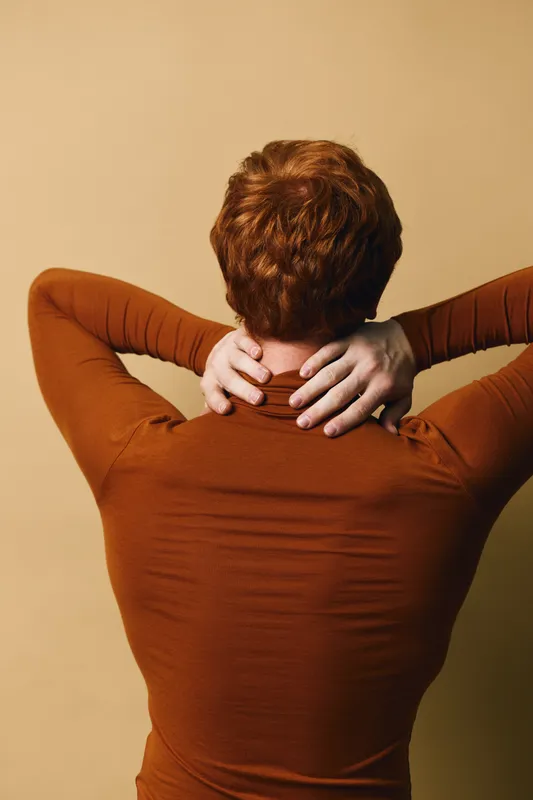 Person with red hair wearing brown long sleeve top holds neck with both hands from behind, standing against plain beige background, showing back and relaxed posture in casual style.