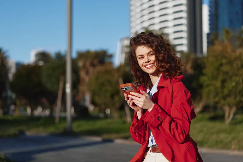 A young woman in a red coat smiles while checking her smartphone in a sunny urban park, with modern buildings and green trees in the background, conveying a casual moment.