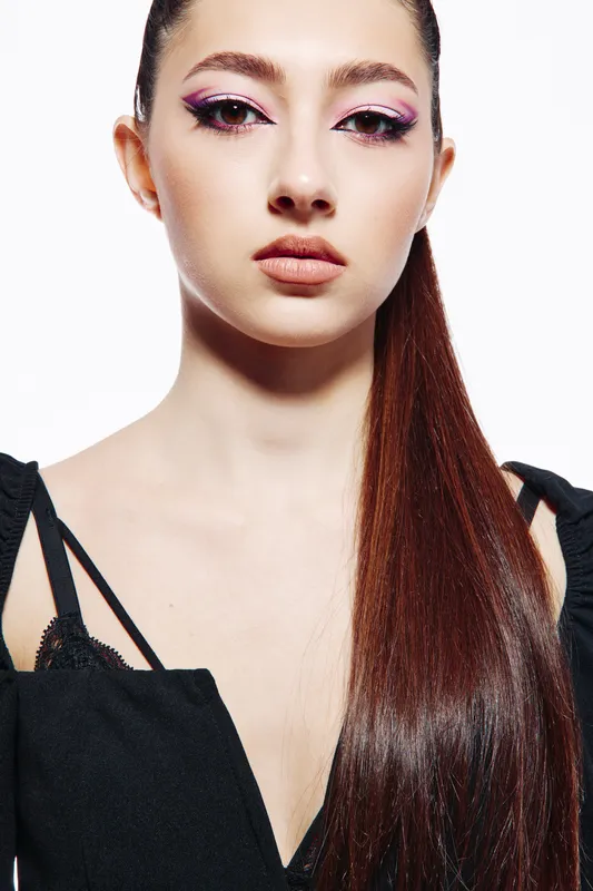 Close-up portrait of a young woman with creative pink eye makeup and long straight hair dressed in a black textured top. Studio lighting emphasizes her smooth skin and beauty.
