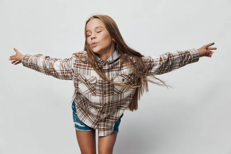 Girl with long hair wearing plaid shirt and denim shorts posing with arms outstretched and eyes closed on white backdrop. Youthful, playful expression with casual style and freedom.