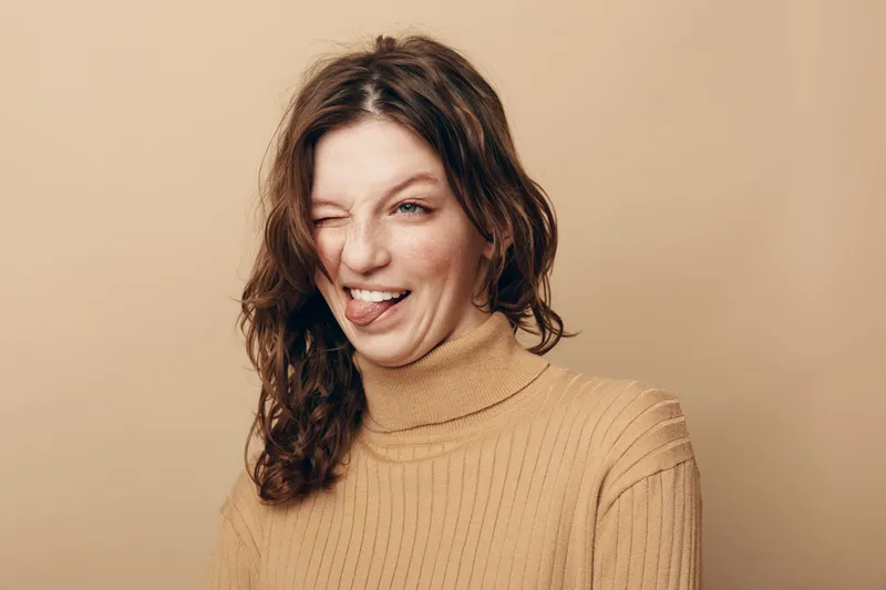Young woman with curly hair in beige sweater makes funny face sticking out tongue and winking. Studio portrait on neutral background with casual style and playful mood.