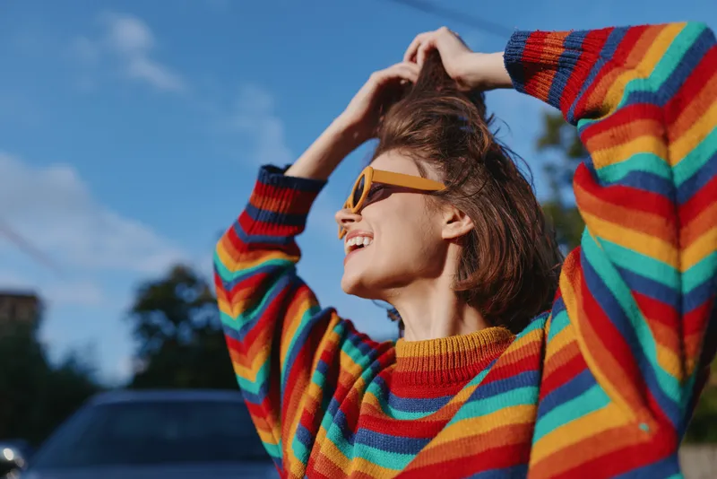 Woman in colorful striped sweater wearing sunglasses, smile and portrait outdoors in sunlight, casual fashion, happy young female enjoying fresh air and relaxed lifestyle.