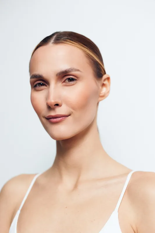 Close up portrait of a confident young woman with flawless skin, subtle makeup, and a calm expression, isolated on a light background, emphasizing natural beauty and skincare for display.