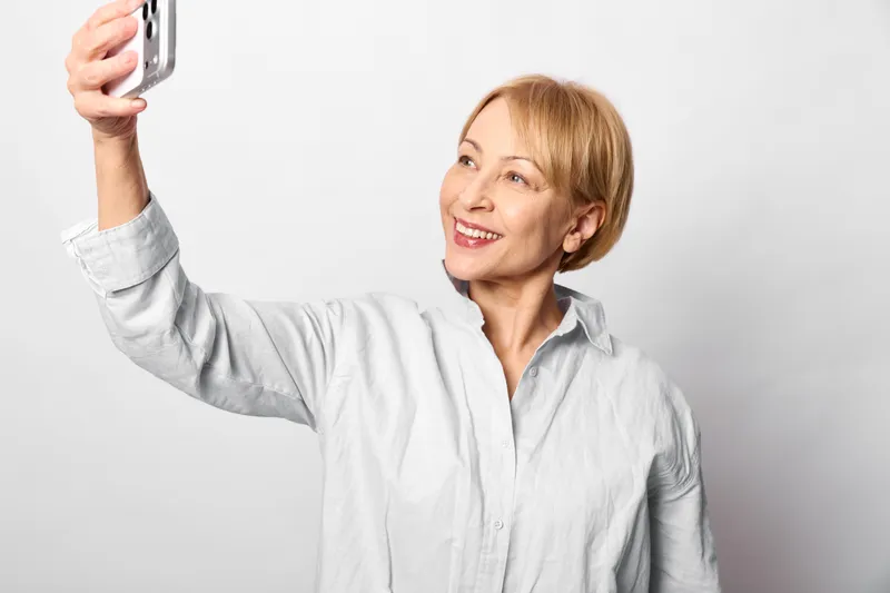 Woman selfie smartphone smiling portrait mature casual indoors white background, senior holds smartphone high for a clean studio selfie, relaxed pose, cheerful eyes, soft white light