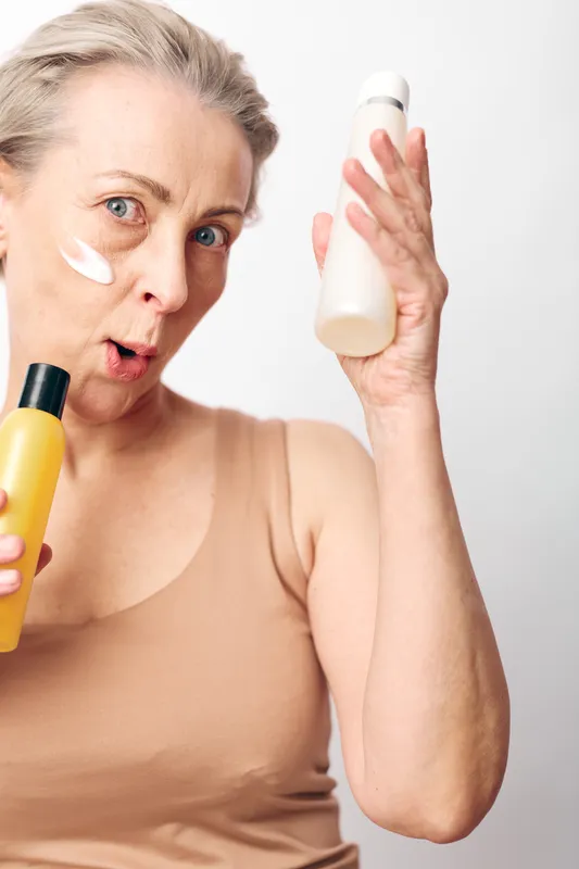 Mature woman with surprised expression holds two skincare bottles and has cream on cheek. Concept of beauty routine, aging skin care, self-care, and natural cosmetic products in bright studio.