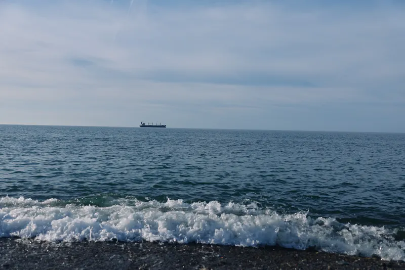 Calm sea with gentle waves crashing on a pebbled shore and a distant ship on the horizon, creating a peaceful seascape under a pale blue sky and soft light reflecting on water.