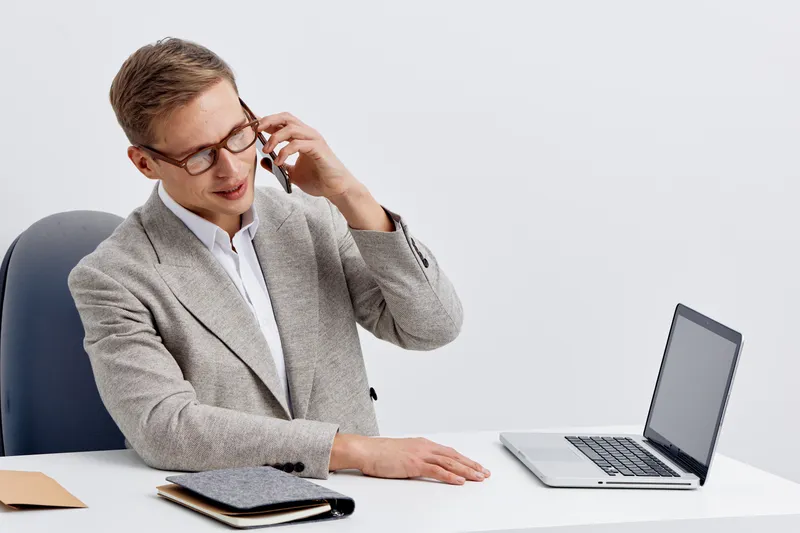 young caucasian man talking smartphone wearing glasses grey blazer sitting office chair laptop digital nomad remote work concept