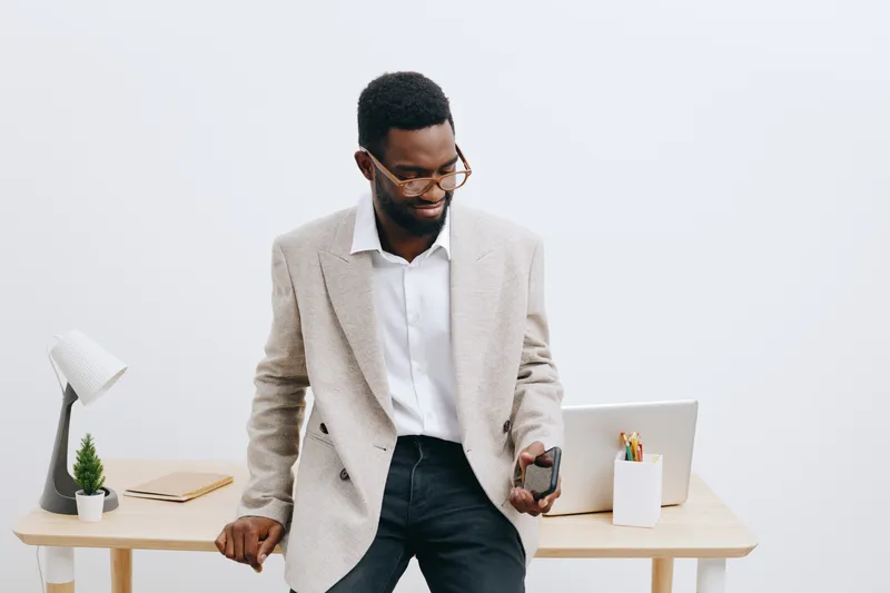 Professional young African American man in business casual attire working on a laptop and holding a smartphone in bright modern office space, concept of work productivity and modern technology.