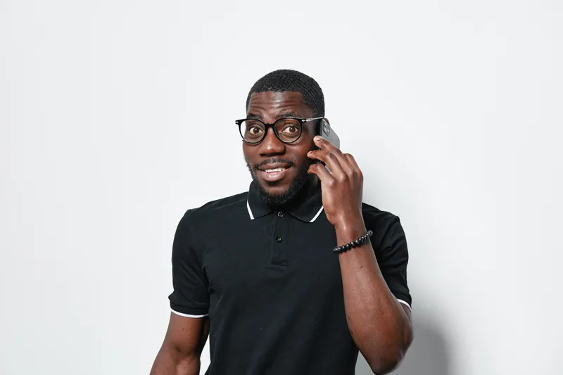 African American man with dark skin wearing glasses and black polo shirt talking on smartphone with smile isolated on white background. Communication, technology and casual style.