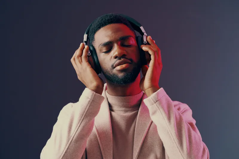 A young Black man enjoying music with headphones, closed eyes, wearing a stylish beige cardigan, set against a colorful gradient background