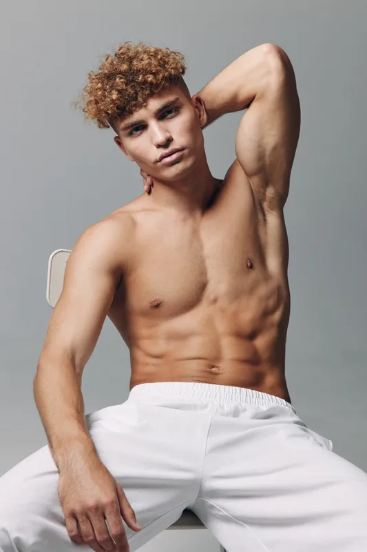 Young muscular man shirtless posing with hand behind head sitting on chair, athletic body with toned abs and curly hair against gray background for fitness and health.