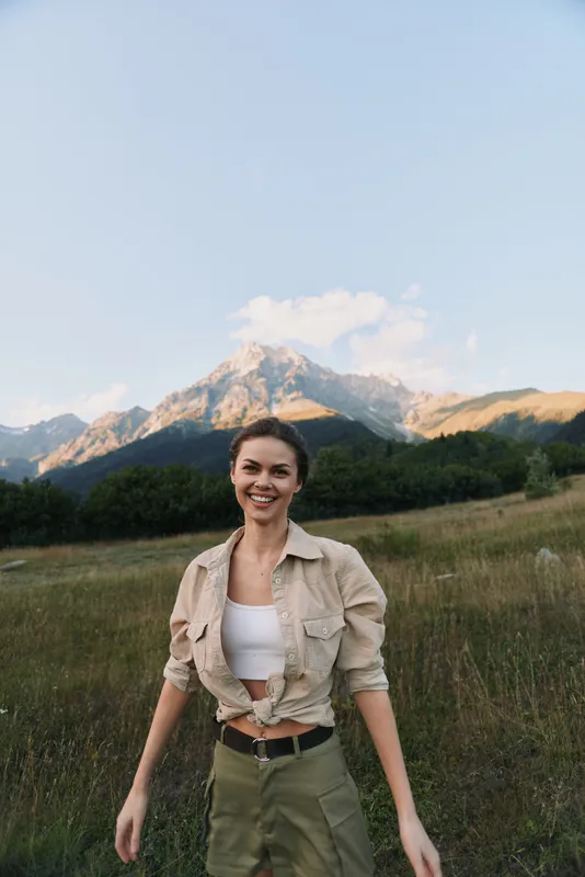 Young woman walks through a sunlit meadow beneath towering mountains, wearing a beige shirt and khaki shorts, smiling with carefree outdoor energy and fresh alpine air.