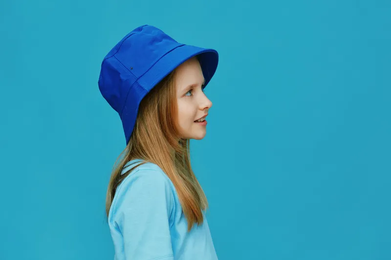 A young girl with long hair wearing a blue bucket hat stands against a vibrant turquoise background, showcasing a joyful expression and a stylish summer look