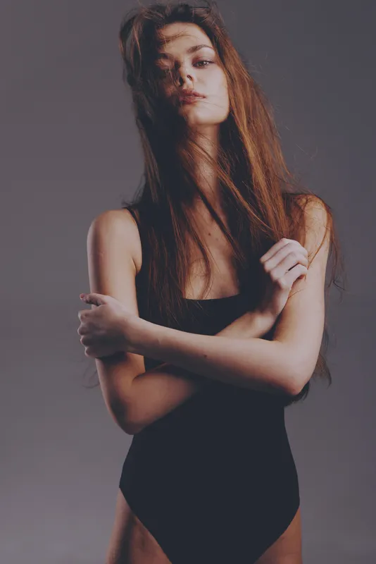 Young woman posing in black bodysuit with confident expression against grey background