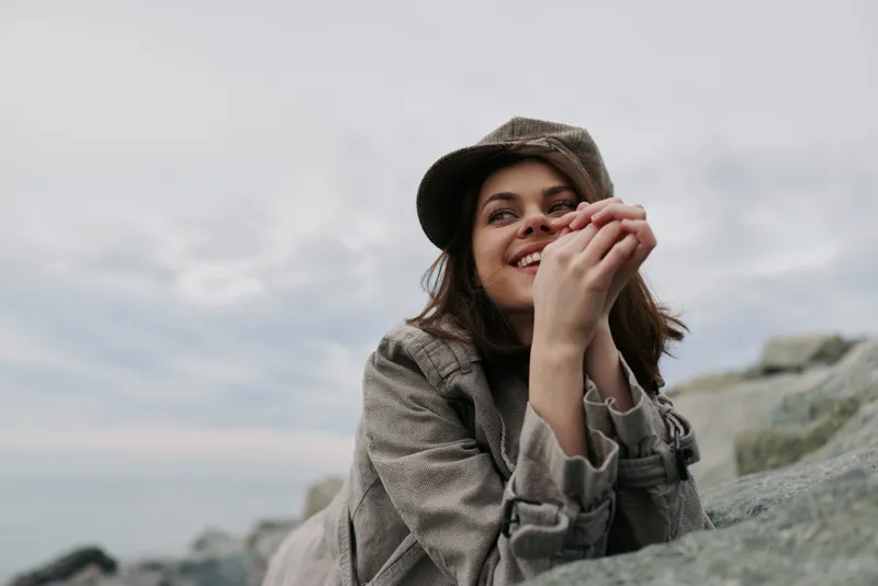 Young woman in a camouflage jacket and hat smiles while resting on rocks by the sea under an overcast sky. Concept for nature, outdoor, relaxation, and happiness.