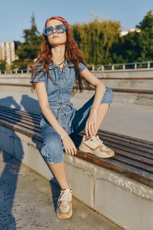 Young woman wearing denim jumpsuit and sunglasses sitting on urban bench with relaxed confident pose under bright sunlight.