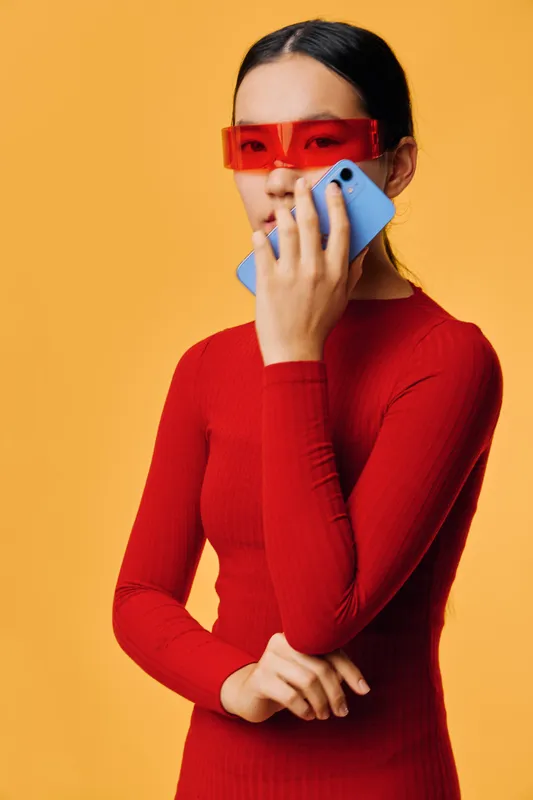 Woman in red dress and futuristic orange glasses holding blue smartphone standing against orange background for modern technology and fashion concept.