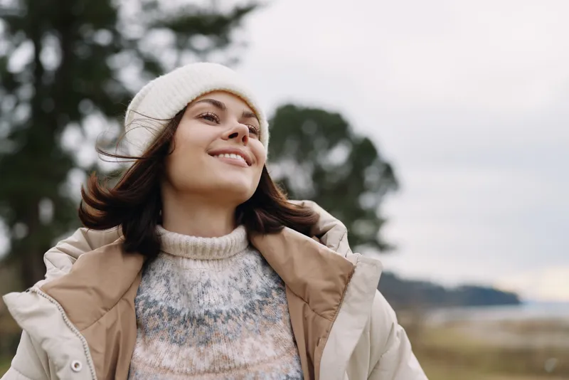Woman outdoors in a cozy beanie and jacket, smiling with the breeze in a natural landscape, capturing warmth, happiness and carefree mood in fresh air.