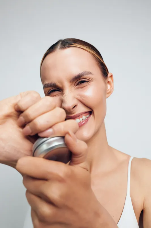 Happy woman smiling with eyes closed, squeezing a metal can opener, close-up portrait, natural skin, casual outfit, bright indoor lighting, light background