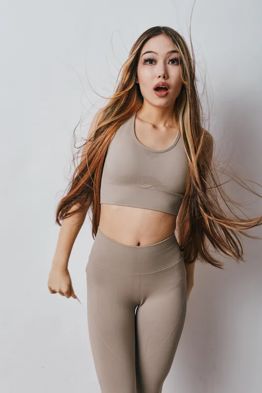Young woman with long hair wearing beige sportswear posing with surprised expression in studio against plain background. Fitness, emotions, active lifestyle and youth concept.