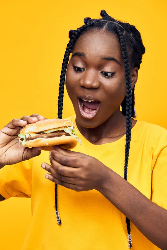 Excited young African woman with braids holding a delicious burger against a vibrant yellow background, showcasing her surprise and joy for fast food