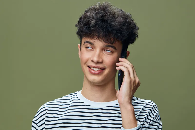 Young man talking on smartphone with a smile, wearing a striped shirt against a muted olive background, casual style and positive expression.