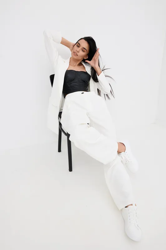 stylish woman sitting in white pants and blazer relaxed on black chair with confident gaze and casual pose in bright minimalist studio setting