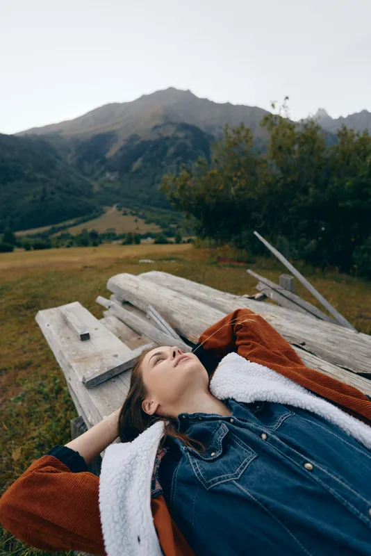 woman meadow mountains bench relaxation nature jacket outdoors portrait of a young woman lying on a rustic bench in a scenic meadow by mountain slopes, peaceful countryside moment of calm and leisure