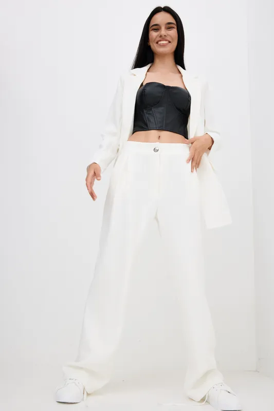 Smiling woman in white wide pants and jacket, wearing black corset top and white sneakers, standing against plain white background with confident posture and natural smile.