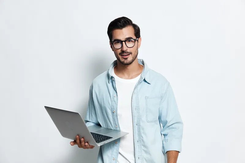 Friendly smiling young man wearing glasses and a light denim shirt, holding a laptop, standing against a clean white background. Technology lifestyle concept.