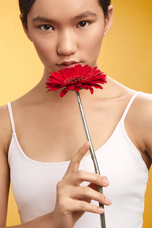 Asian woman gently holds a red flower near her lips, creating a soft, intimate portrait. The warm background highlights her calm demeanor and delicate features, evoking elegance and quiet confidence.
