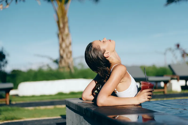 Young woman in white swimsuit relaxes by poolside leaning on edge holds cold drink enjoys sunny summer day with eyes closed outdoors in tropical lifestyle setting.