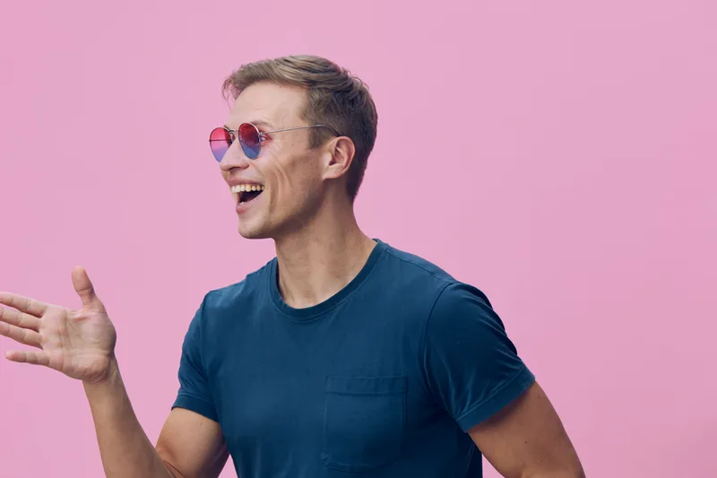 Smiling man wearing colorful heart-shaped sunglasses cheerful expression casual blue t-shirt pink background studio portrait. People lifestyle concept