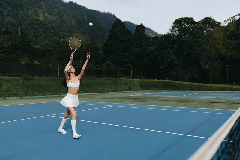 Young athletic woman in a tennis outfit serving on a blue court, showcasing energy and determination amidst a natural green backdrop
