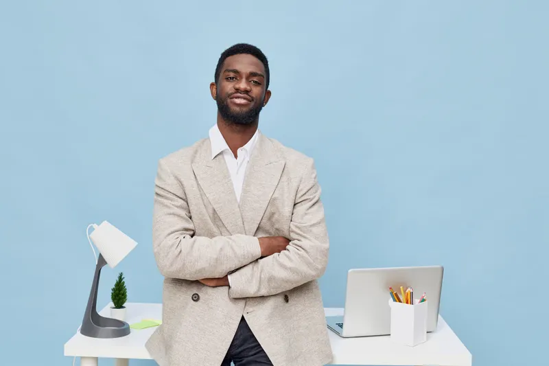 confident man smiling young African American businessman wearing beige blazer standing at modern office desk isolated on light blue background. Corporate, professional, success, technology, workspace,