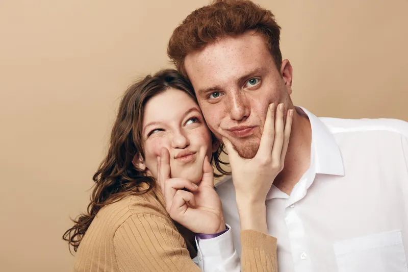 Young woman playfully pinches young man’s cheeks while both smile and pose against beige background. Concept of friendship, fun, affection, and casual lifestyle in studio portrait.