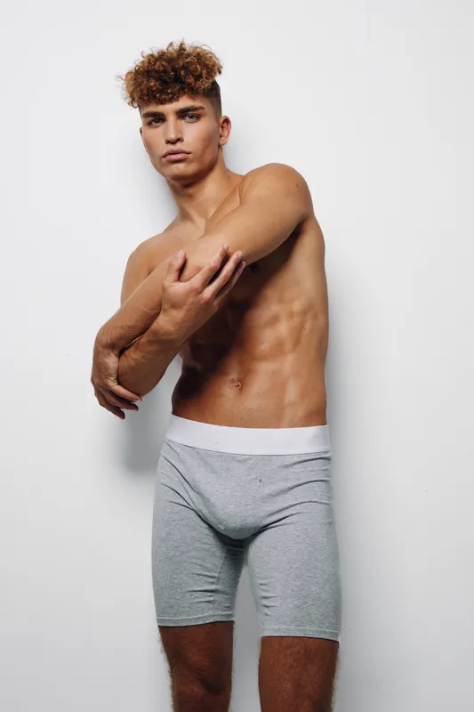 Young man with curly hair posing shirtless in gray boxer briefs against white background, showing fit body and confident expression in studio lighting.