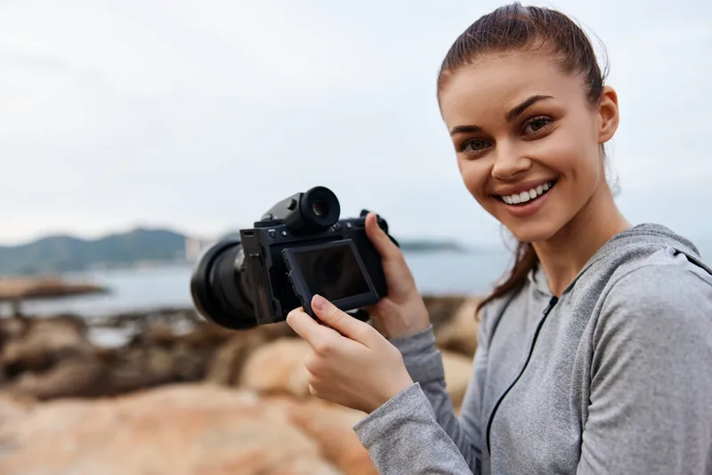 Happy young woman with camera on rocky beach, enjoying photography during sunset, showcasing passion for nature and adventure, vibrant colors and natural scenery