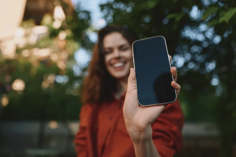 A smiling woman in casual attire holds a phone toward the camera in an outdoor park, capturing a candid lifestyle moment with bright light, greenery, and a friendly atmosphere, showcasing modern tech