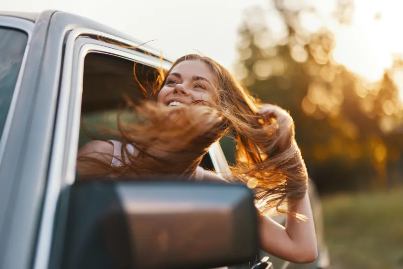 Happy young woman with flowing hair enjoying a road trip in nature, sunlit background, carefree emotion, vibrant colors, summer adventure concept.