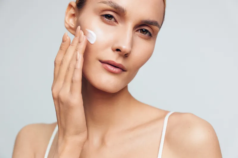 A young woman with clear skin applies moisturizing cream to her face with her hand against a neutral background. Concept for skincare, beauty routine, and healthy skin care.