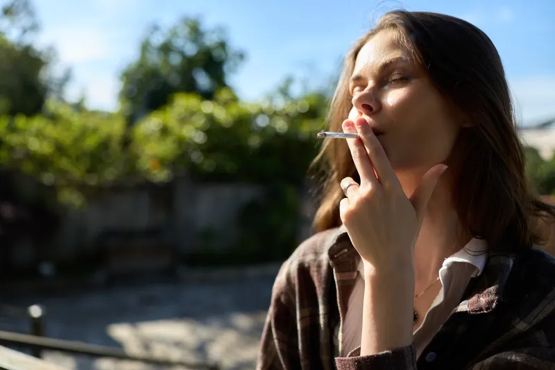 A woman outdoors smokes a cigarette in bright sunlight, casual jacket, relaxed mood. A lifestyle moment depicting personal routine, coping, and quiet reflection