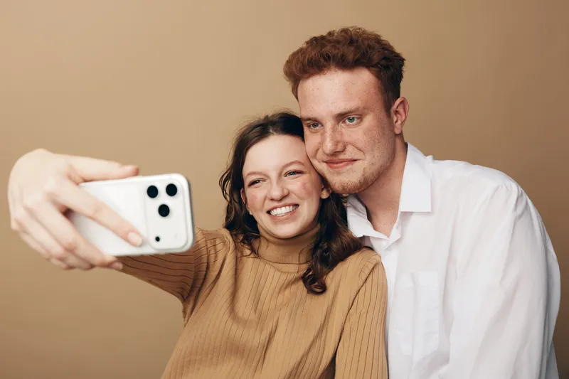 Smiling young couple takes selfie with smartphone on beige background. Happy man and woman enjoy moment together, casual clothing, natural light, concept of friendship and modern technology.