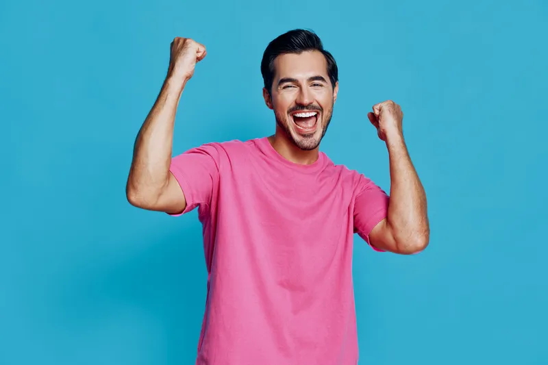 Cheerful happy young man in pink t-shirt raising hands with excitement celebrating success against solid blue background. Glowing positivity and confidence concept.