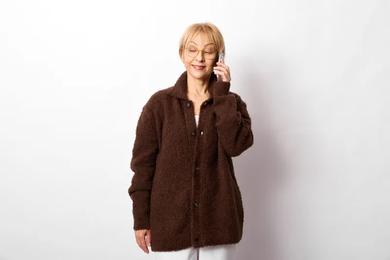 Woman with glasses phone coat portrait, indoor smiling mature blonde, warm brown fur coat, holding phone to ear, relaxed pose, soft studio light, white background
