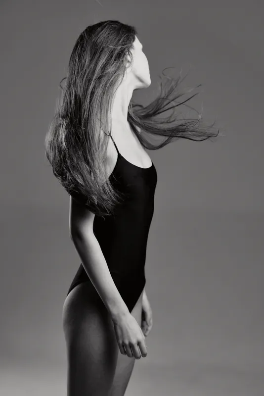 Black and white portrait of a woman with flowing hair wearing a black swimsuit on gray background.