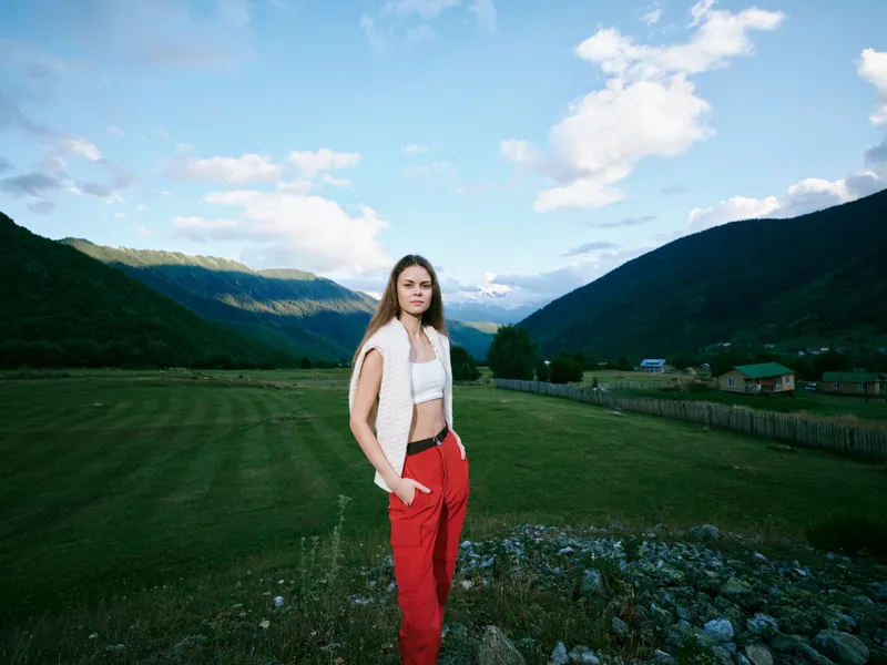Woman standing in a green countryside field with distant mountains, wearing a white crop top and red pants, confident pose under bright sky, vivid landscape and natural light.